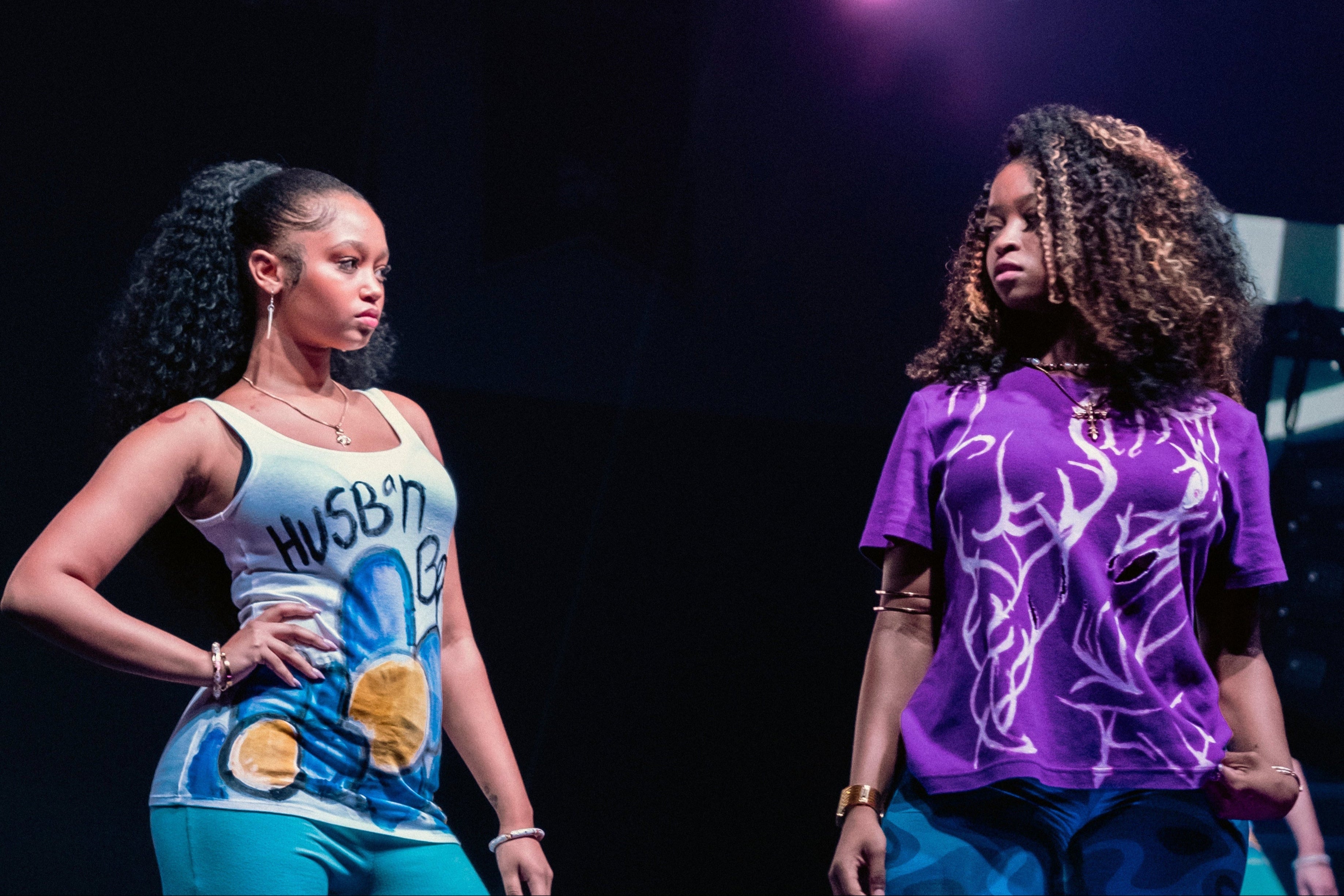 Two women on stage with one wearing a blue and white tank top and the other in a purple shirt with a white design.